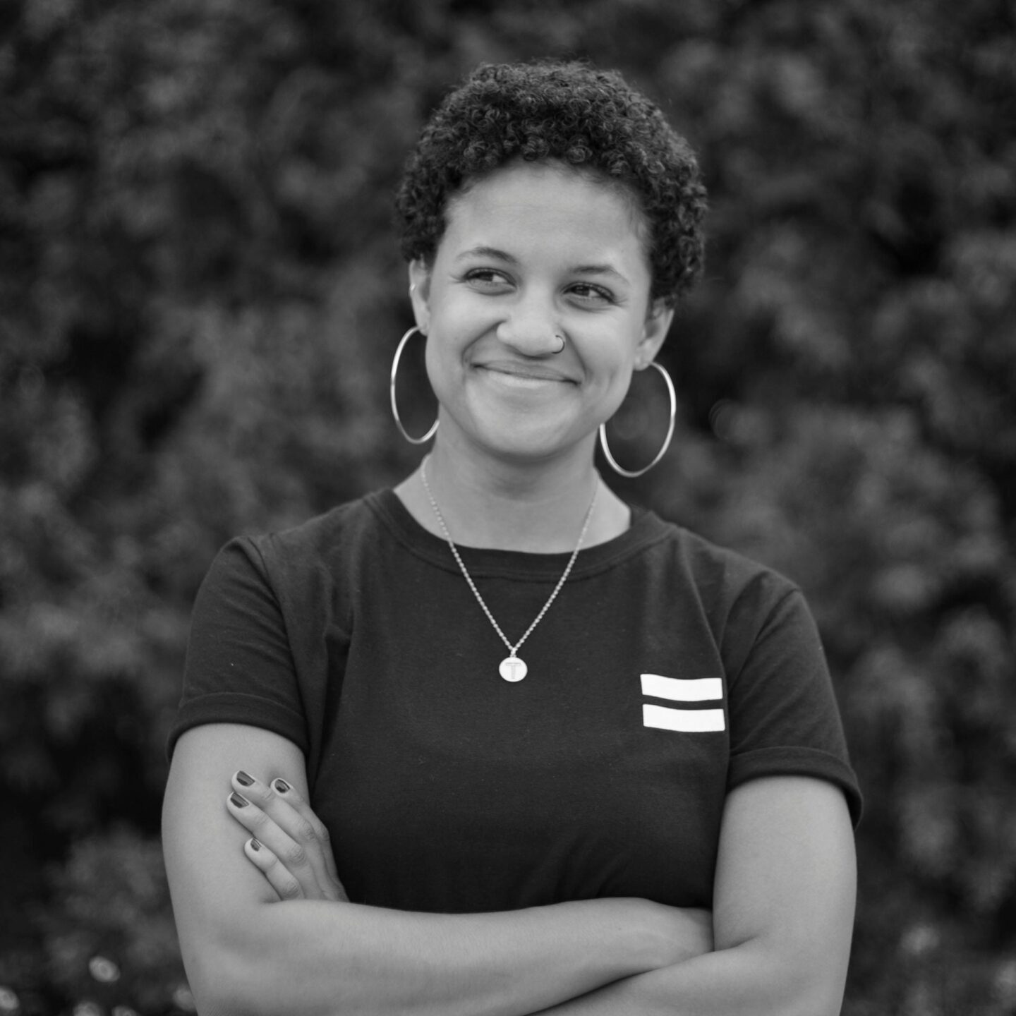 Portrait of a smiling woman with hoop earrings and curly hair, arms crossed, captured in black and white.